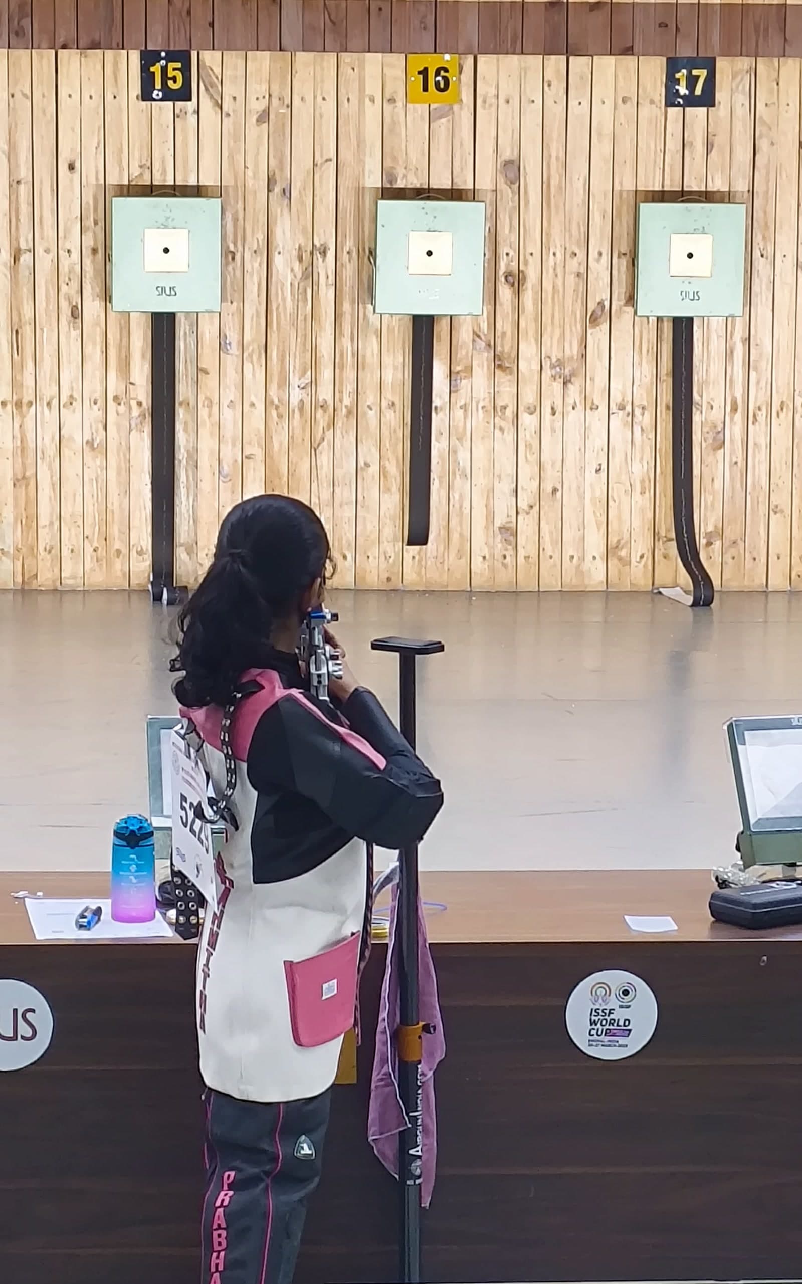Brahmitha G at the firing line, seen from behind, aiming her air rifle at a row of electronic targets