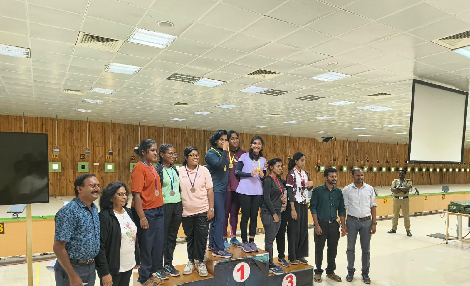 Medal ceremony at an indoor range, with three teams on the podium and Brahmitha G on the top step